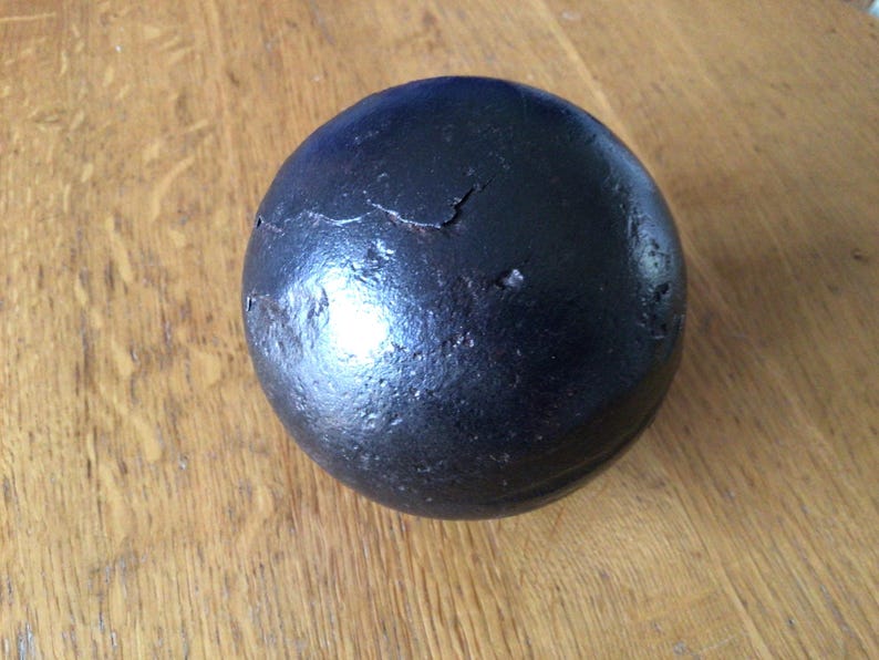 May include: A dark, spherical object with a textured surface and visible cracks. The object is a solid black colour and appears to be made of metal. It sits on a wooden surface.
