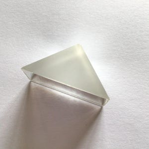 Puede incluir: Un prisma triangular transparente de vidrio. El prisma está sobre una superficie blanca y proyecta una sombra. La forma es un triángulo rectángulo con un aspecto esmerilado en la superficie superior.