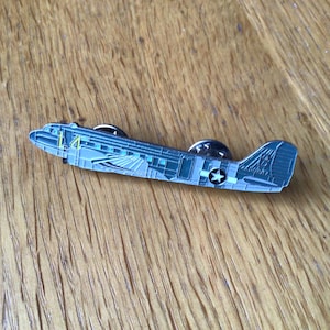 May include: A detailed metal airplane pin in shades of grey and blue, featuring a star emblem and the number 14. The pin is set against a wooden background, showcasing the intricate design of the aircraft.