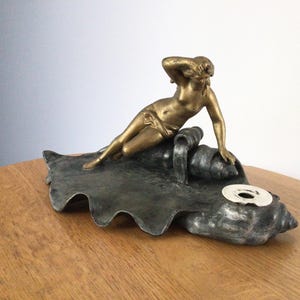 May include: A metallic sculpture featuring a gold-toned figure reclining on a dark, textured, wave-like base. The figure rests one arm on a large, dark shell-shaped element that includes a circular white insert with a central hole.