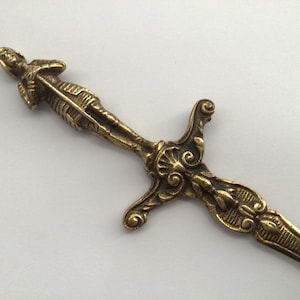 May include: A detailed, antique-style letter opener in a bronze finish. The handle features a sculpted figure, and the blade has an ornate design. The overall design is reminiscent of a small sword.