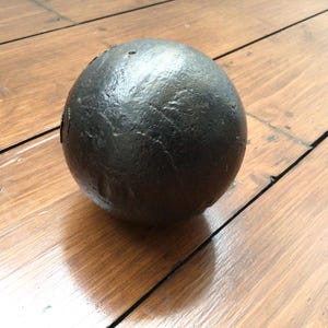 May include: A dark grey sphere with a textured surface, resting on a wooden surface. The ball's surface has a rough texture. The wooden surface has visible wood grain. The sphere appears to be made of metal.