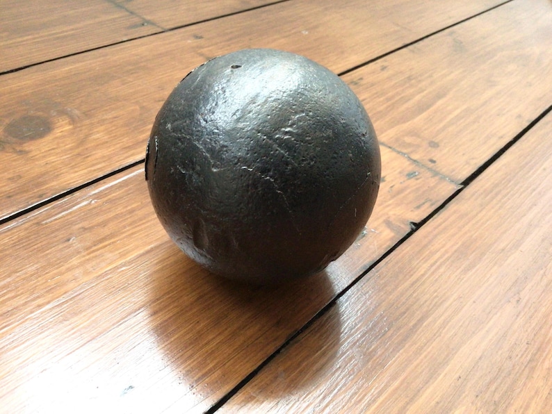 May include: A dark grey sphere with a textured surface, resting on a wooden surface. The ball's surface has a rough texture. The wooden surface has visible wood grain. The sphere appears to be made of metal.