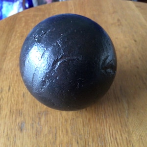 May include: A dark grey sphere with a textured surface. The sphere has an irregular shape and appears to be made of a dense material. It rests on a wooden surface.