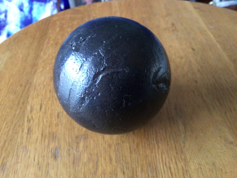 May include: A dark grey sphere with a textured surface. The sphere has an irregular shape and appears to be made of a dense material. It rests on a wooden surface.