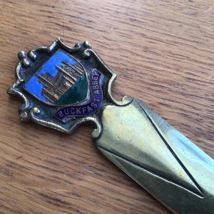 Könnte beinhalten: Antiker Brieföffner aus Messing mit einem dekorativen Schild, das das blaue und grüne Emblem der Buckfast Abbey zeigt. Der Öffner hat eine spitze Klinge und einen detaillierten Griff.