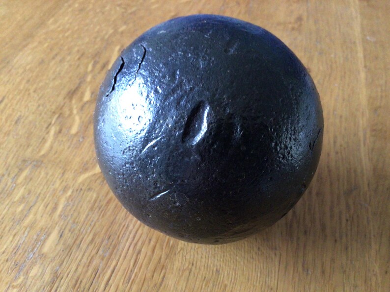 May include: A dark, round, solid black object with a textured surface and visible cracks. The object is spherical and appears to be made of a dense material. The background is a wooden surface.
