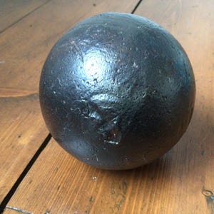 May include: A dark, spherical object with a textured surface, possibly metal. It sits on a wooden surface with visible grain. The sphere appears solid and heavy. The object's diameter is approximately 10 cm.