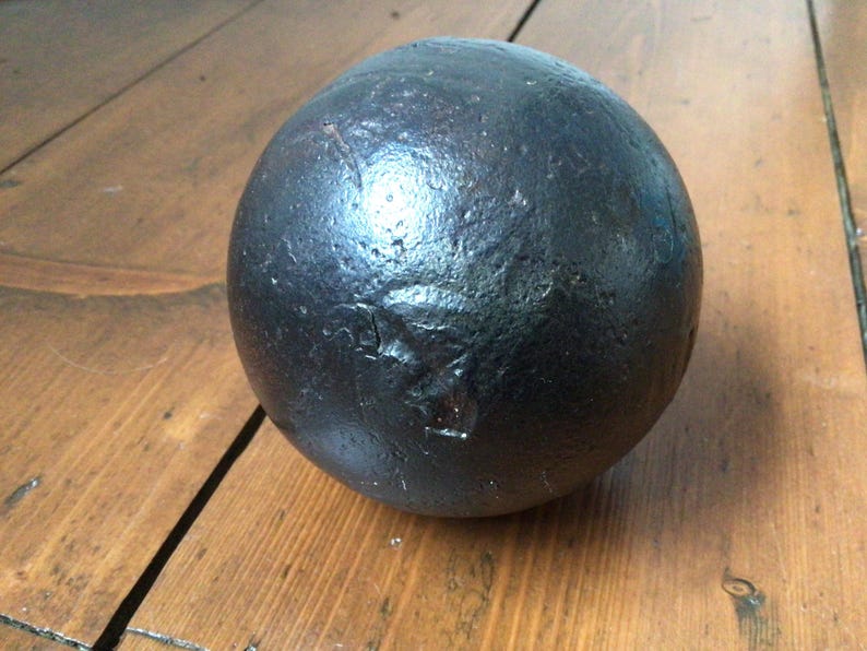 May include: A dark, spherical object with a textured surface, possibly metal. It sits on a wooden surface with visible grain. The sphere appears solid and heavy. The object's diameter is approximately 10 cm.