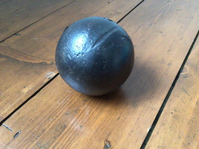 May include: A dark, spherical object with a textured surface, possibly metal. The ball has a visible seam and rests on a wooden surface with visible grain.