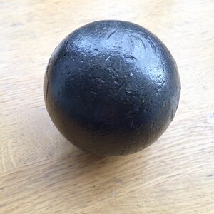 May include: A black sphere with a textured surface, set against a light-coloured wooden background. The sphere's material is likely heavy, possibly metal or stone. The design is simple and industrial, with a focus on the object's form.