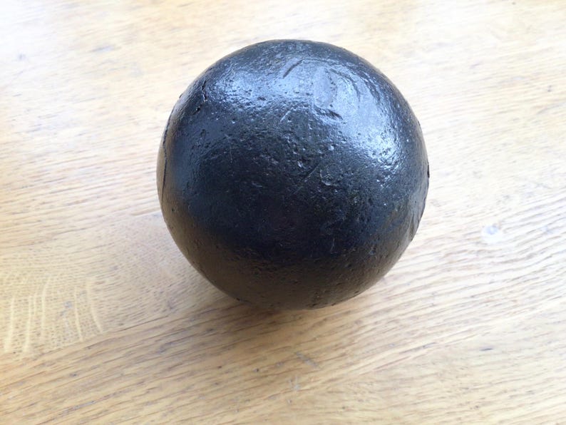 May include: A black sphere with a textured surface, set against a light-coloured wooden background. The sphere's material is likely heavy, possibly metal or stone. The design is simple and industrial, with a focus on the object's form.