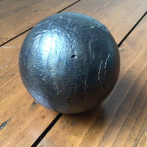 May include: A dark grey, spherical object with a textured surface. The ball appears to be made of metal and has some visible imperfections. It rests on a wooden surface with visible wood grain.