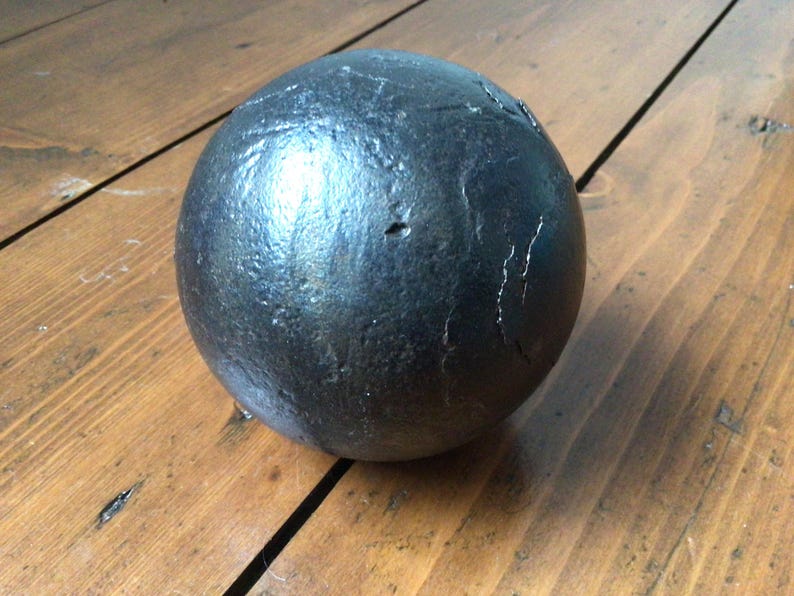 May include: A dark grey, spherical object with a textured surface. The ball appears to be made of metal and has some visible imperfections. It rests on a wooden surface with visible wood grain.