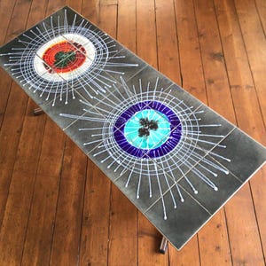 May include: Rectangular coffee table with a dark gray tiled surface. The tiles feature a colorful, abstract design with concentric circles in red, white, blue, and turquoise. The table has chrome legs and is set on a wooden floor.