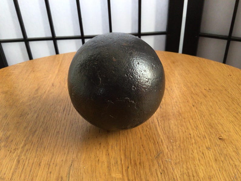 May include: A dark, spherical object with a textured surface, possibly metal, on a wooden table. The sphere is black and has a slightly reflective sheen. The background features a window and a black lattice.