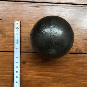 May include: A dark, round object with a textured surface, possibly metal, rests on a wooden surface. A ruler is placed beside the object, showing measurements in inches. The object is approximately 10 cm in diameter.