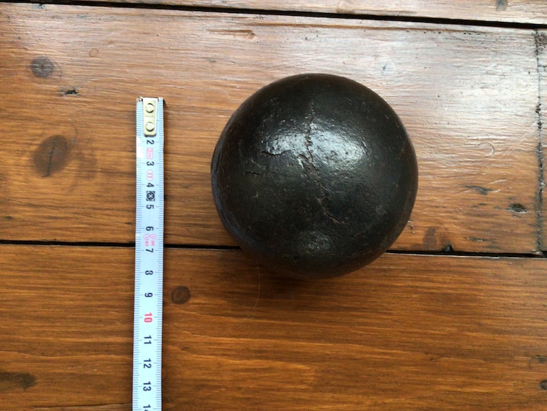 May include: A dark, round object with a textured surface, possibly metal, rests on a wooden surface. A ruler is placed beside the object, showing measurements in inches. The object is approximately 10 cm in diameter.