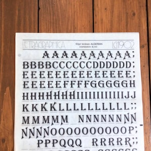 Puede incluir: Una hoja blanca de letras de transferencia en seco Letragraphica con la tipografía Algerian en tamaño 60pt 14.6mm. La hoja muestra varias filas de letras mayúsculas negras de la A a la S, incluyendo signos de puntuación, con el texto LG1902 visible.