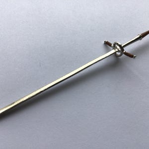 May include: A miniature silver sword with a brown handle and crossguard. The sword features a decorative ring near the hilt. The blade is long and slender, and the overall design is detailed and elegant.