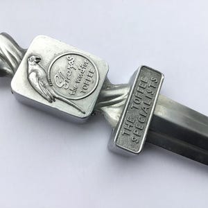 Puede incluir: Un martillo para toffee de color plateado con una cabeza rectangular con un pájaro y las palabras "Sharps the Toffee". El mango dice "The Toffee Specialists".