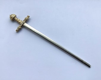 May include: A decorative letter opener shaped like a sword. The handle is gold-colored with intricate detailing, and the blade is silver. The letter opener is lying on a white surface.