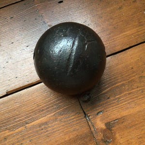 May include: A dark grey cannonball with a textured surface and a vertical indentation. The spherical object is displayed on a wooden surface with visible wood grain. The cannonball is a historical item.