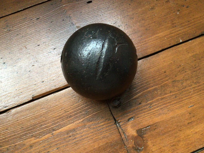 May include: A dark grey cannonball with a textured surface and a vertical indentation. The spherical object is displayed on a wooden surface with visible wood grain. The cannonball is a historical item.