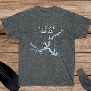 May include: Dark gray t-shirt with a light blue map of a lake and the words "SINCLAIR Lake Life". The shirt is on a wooden surface with a paddle, rope, and flip-flops.