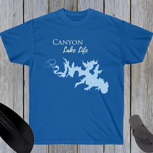 May include: A blue t-shirt with the text "Canyon Lake Life" and a light blue lake outline graphic. The shirt is displayed on a wooden surface with a paddle, rope, and flip-flops.