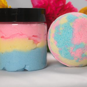 May include: A clear plastic jar filled with layered pink, yellow, and blue whipped body butter topped with a black lid, positioned next to a spherical bath bomb featuring a marbled pattern of pink, yellow, and light blue colors.