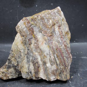 Puede incluir: Una muestra de roca en bruto con una apariencia estratificada. La piedra presenta bandas de tonos blancos, marrones y rojizos. La superficie de la roca es texturizada e irregular, con una mezcla de áreas opacas y translúcidas. La forma general es angular y asimétrica.