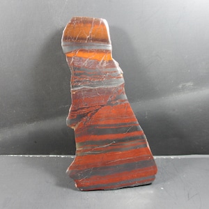 Pode incluir: Uma pedra polida, de forma irregular, com faixas vermelhas, laranja e cinza. A pedra tem uma forma natural e orgânica e uma superfície lisa. As variações de cor criam um efeito visual marcante, adequado para exibição ou coleção.