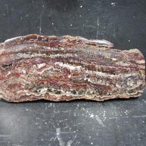 May include: A rectangular, layered stone specimen with bands of red, brown, white, and gray. The natural rock formation has a rough, textured surface and is displayed against a dark background. The stone appears to be a type of agate or similar mineral.
