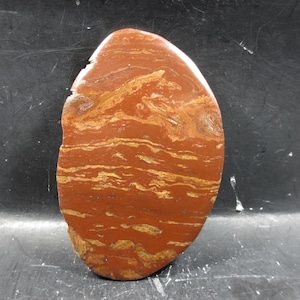 Puede incluir: Una piedra pulida, de forma ovalada, con un color base marrón rojizo y patrones en capas de tonos dorados y beige. La piedra tiene una forma y textura natural y orgánica, mostrando sus características geológicas únicas.