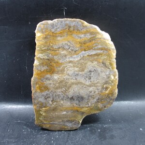 Puede incluir: Una losa de piedra natural con un patrón único de colores en capas. La piedra presenta bandas de tonos grises, blancos y amarillo dorado. La forma irregular y la superficie pulida resaltan su belleza natural.