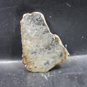 Puede incluir: Una piedra natural de forma irregular con un patrón veteado gris claro y blanco. La piedra tiene detalles en gris oscuro y marrón y un borde rugoso y texturizado. La piedra se muestra sobre un fondo oscuro.