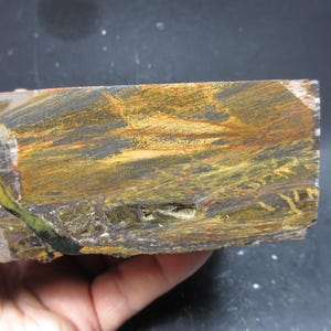 Puede incluir: Un trozo rectangular de piedra ojo de tigre, que muestra una mezcla de tonos amarillo dorado, marrón y negro. La superficie de la piedra exhibe patrones y texturas naturales, con capas y variaciones de color visibles.