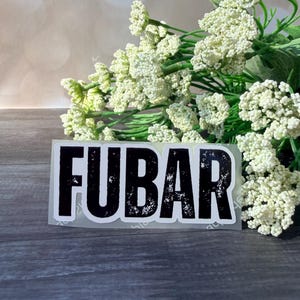 Puede incluir: Una pegatina blanca con la palabra "FUBAR" en letras negras en negrita. La pegatina está colocada sobre una superficie de madera oscura, con un ramo de flores blancas en el fondo.