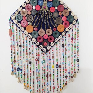 K&ouml;nnte beinhalten: Ein rautenf&ouml;rmiger Wandbehang mit einem blauen Samt-Hintergrund, der mit bunten Kn&ouml;pfen, Muscheln und Perlen verziert ist. Der Behang hat ein zentrales Design aus goldenen Ketten und einem rosafarbenen Juwel. Der untere Teil des Behangs besteht aus mehreren Str&auml;ngen bunter Perlen mit blauen Gl&uuml;cksbringer-Augen.