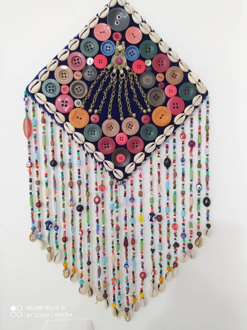 K&ouml;nnte beinhalten: Ein rautenf&ouml;rmiger Wandbehang mit einem blauen Samt-Hintergrund, der mit bunten Kn&ouml;pfen, Muscheln und Perlen verziert ist. Der Behang hat ein zentrales Design aus goldenen Ketten und einem rosafarbenen Juwel. Der untere Teil des Behangs besteht aus mehreren Str&auml;ngen bunter Perlen mit blauen Gl&uuml;cksbringer-Augen.