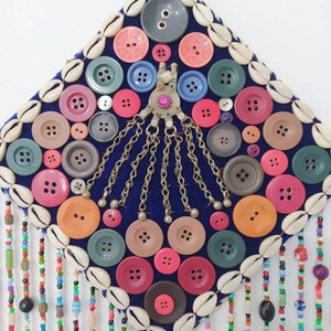 K&ouml;nnte beinhalten: Ein quadratischer Wandbehang aus blauem Samt, verziert mit bunten Kn&ouml;pfen, Muscheln und Perlenfransen. Die Kn&ouml;pfe sind in einem Kreis um ein zentrales Metallst&uuml;ck mit Ketten angeordnet.