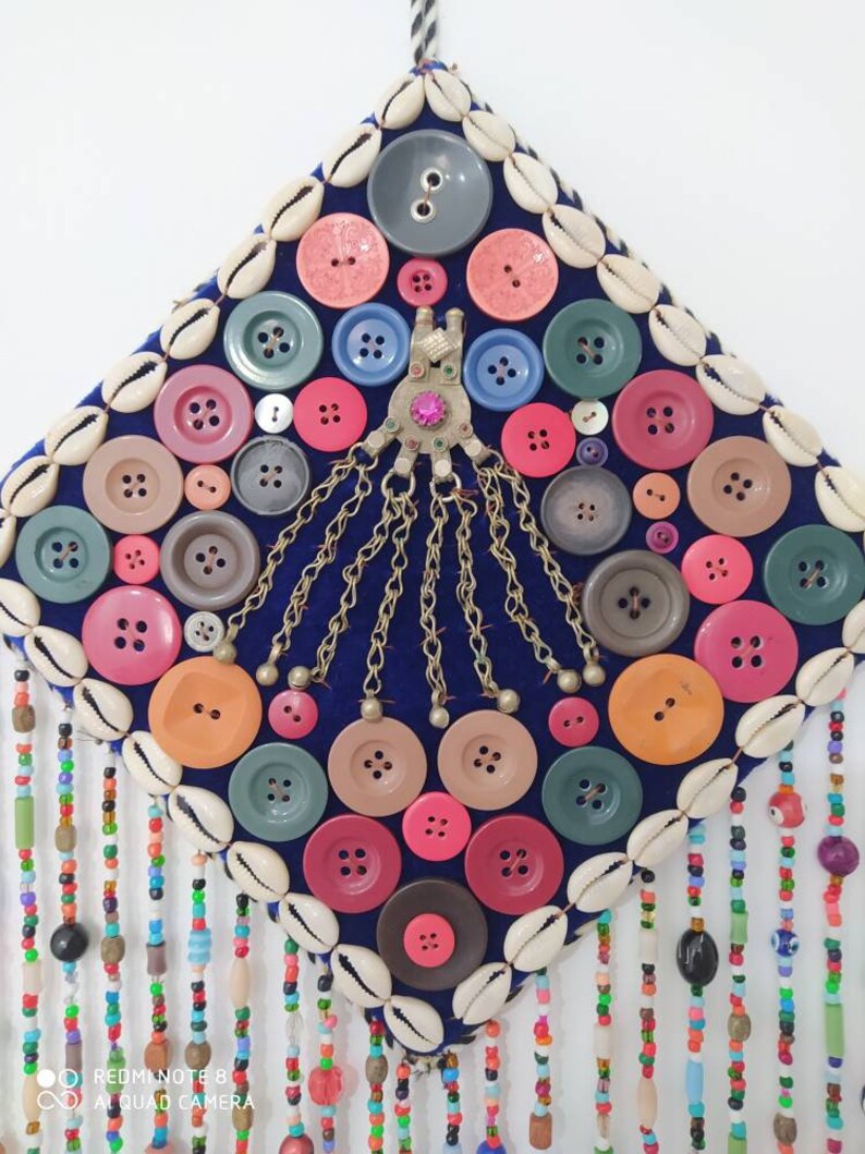 K&ouml;nnte beinhalten: Ein quadratischer Wandbehang aus blauem Samt, verziert mit bunten Kn&ouml;pfen, Muscheln und Perlenfransen. Die Kn&ouml;pfe sind in einem Kreis um ein zentrales Metallst&uuml;ck mit Ketten angeordnet.