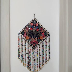 K&ouml;nnte beinhalten: Ein farbenfroher Wandbehang mit einem rautenf&ouml;rmigen Zentrum aus Stoff und Kn&ouml;pfen. Das Zentrum ist von einem Rand aus Muscheln umgeben. Der Behang hat lange F&auml;den aus Perlen und Muscheln, die von unten h&auml;ngen.