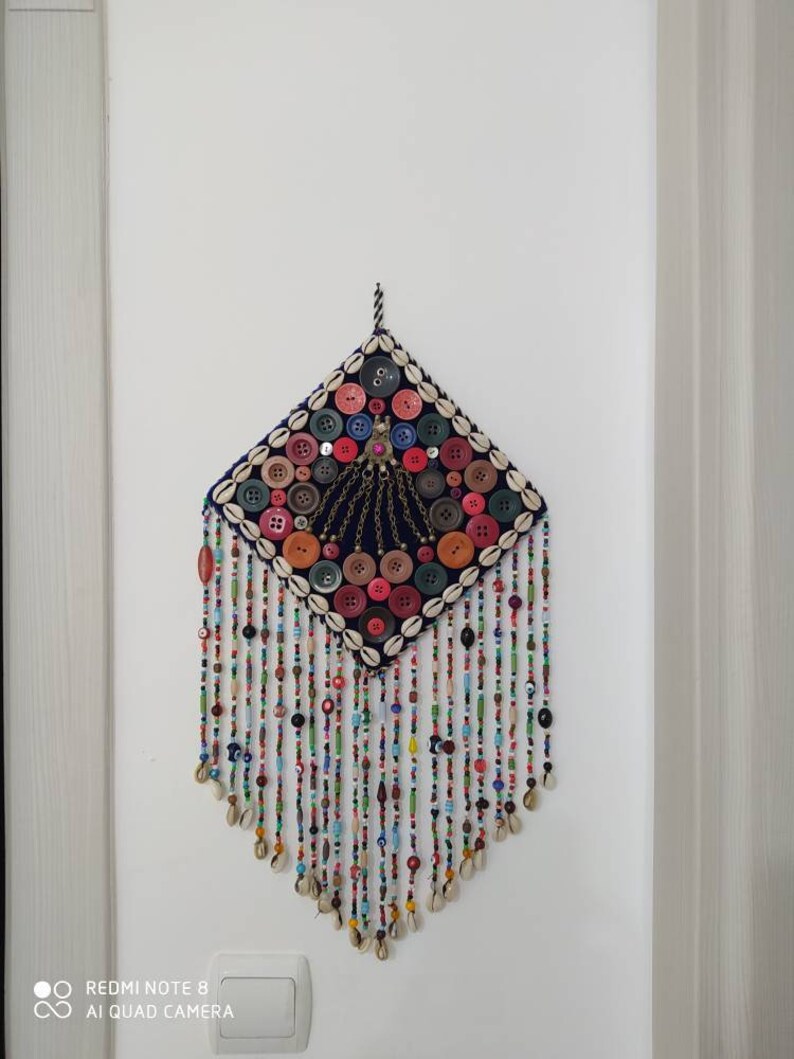 K&ouml;nnte beinhalten: Ein farbenfroher Wandbehang mit einem rautenf&ouml;rmigen Zentrum aus Stoff und Kn&ouml;pfen. Das Zentrum ist von einem Rand aus Muscheln umgeben. Der Behang hat lange F&auml;den aus Perlen und Muscheln, die von unten h&auml;ngen.