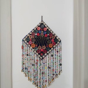K&ouml;nnte beinhalten: Ein farbenfroher Wandbehang mit quadratischer Form und einem Fransenbesatz aus Perlen. Die Mitte des Behangs ist mit Kn&ouml;pfen, Muscheln und Perlen in verschiedenen Farben verziert. Der Behang ist mit einer Schnur an einer wei&szlig;en Wand befestigt.