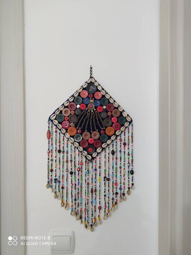 K&ouml;nnte beinhalten: Ein farbenfroher Wandbehang mit quadratischer Form und einem Fransenbesatz aus Perlen. Die Mitte des Behangs ist mit Kn&ouml;pfen, Muscheln und Perlen in verschiedenen Farben verziert. Der Behang ist mit einer Schnur an einer wei&szlig;en Wand befestigt.