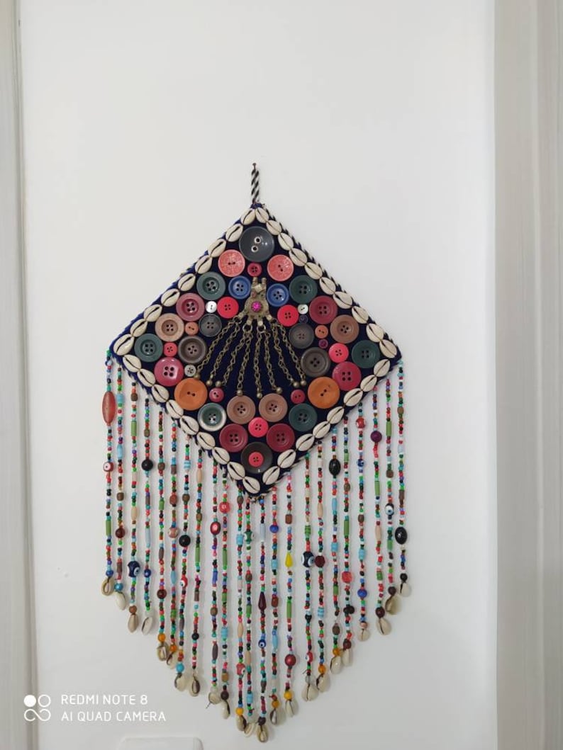 K&ouml;nnte beinhalten: Ein farbenfroher Wandbehang mit einer rautenf&ouml;rmigen Stoffbasis, die mit Kn&ouml;pfen und Muscheln bedeckt ist. Die Basis ist von Str&auml;ngen bunter Perlen mit blauen Gl&uuml;cksaugen umgeben.