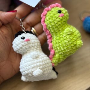 Pode incluir: Dois chaveiros de dinossauro de crochê. Um é branco com detalhes em preto e uma bochecha rosa. O outro é verde limão com detalhes em rosa e um olho preto. Ambos têm argolas prateadas.