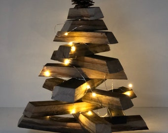 May include: A handcrafted wooden Christmas tree with a natural wood finish. The tree is constructed from stacked rectangular blocks and adorned with a pinecone at the top. Tiny string lights are wrapped around the tree, illuminating the wooden structure.